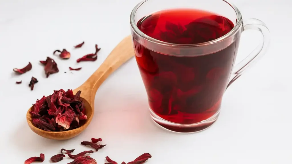 Chá de hibisco em uma xícara de vidro transparente sobre uma mesa branca, ao lado de uma colher de madeira com pétalas secas de hibisco e flores espalhadas ao redor.